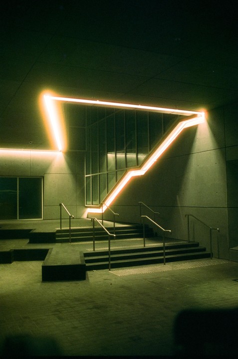An illuminated orange neon light in the shape of a triangle is mounted on a concrete wall above a set of stairs with metal railings.