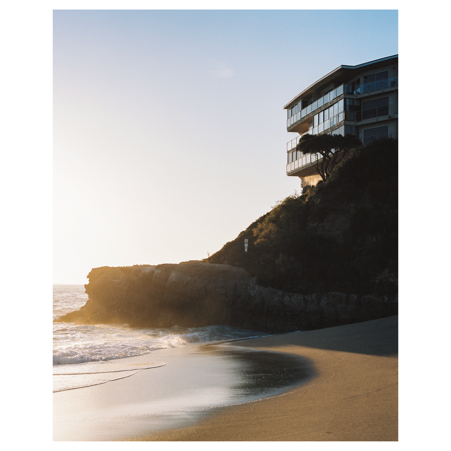 A modern building with balconies sits atop a cliff overlooking a beach with waves.