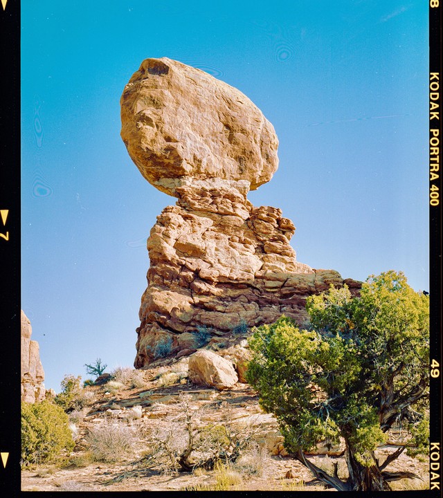 A large balanced rock formation stands against a clear blue sky, surrounded by desert vegetation.