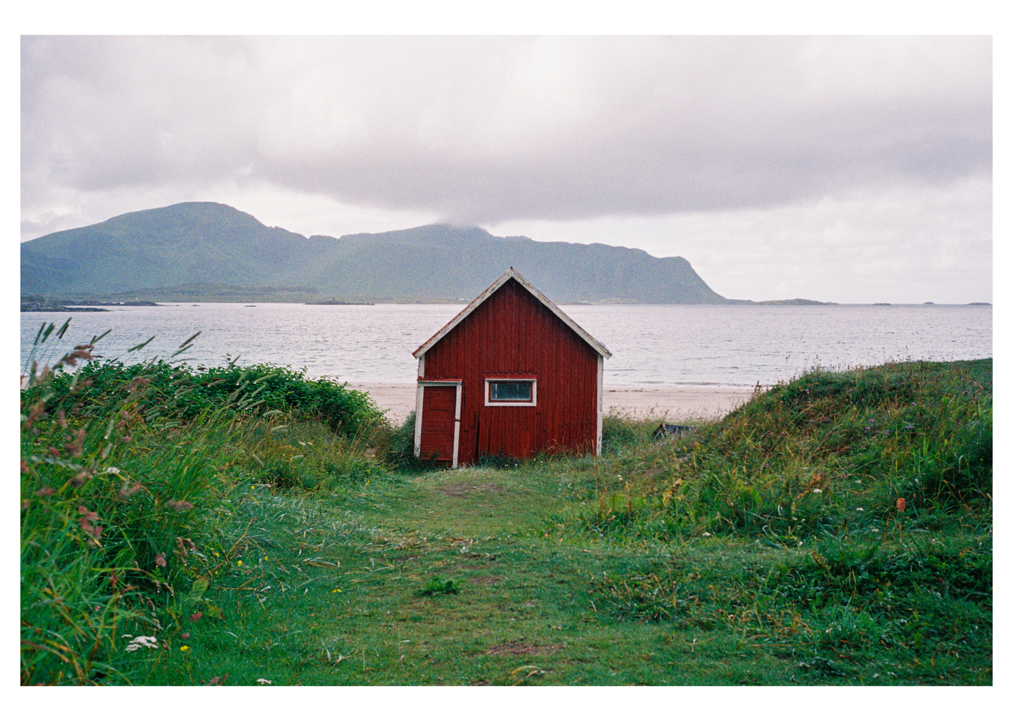 A red cabin sits on a grassy dune overlooking a beach and the sea with mountains in the distance under a cloudy sky.