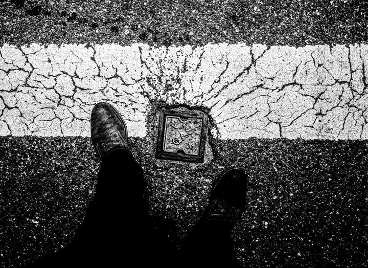 A pair of feet stand on a cracked asphalt road with a white line and a metal plate.