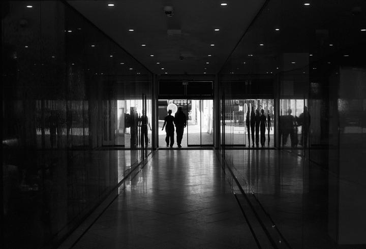 Two people walk through a brightly lit doorway, silhouetted against the light, with other people visible in the reflections of the glass walls.