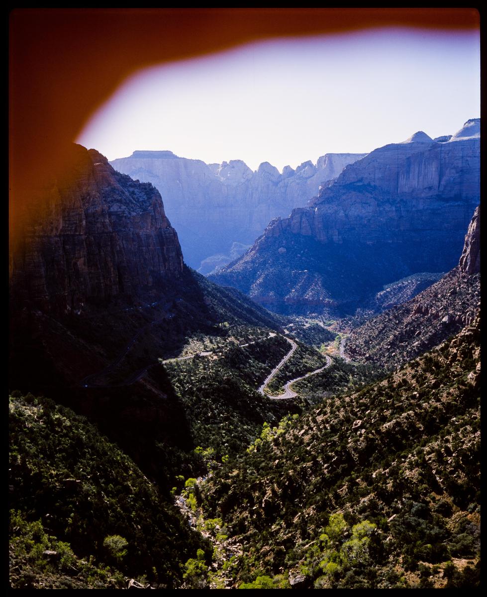 A winding road snakes through a deep valley surrounded by towering mountains under a bright sky.