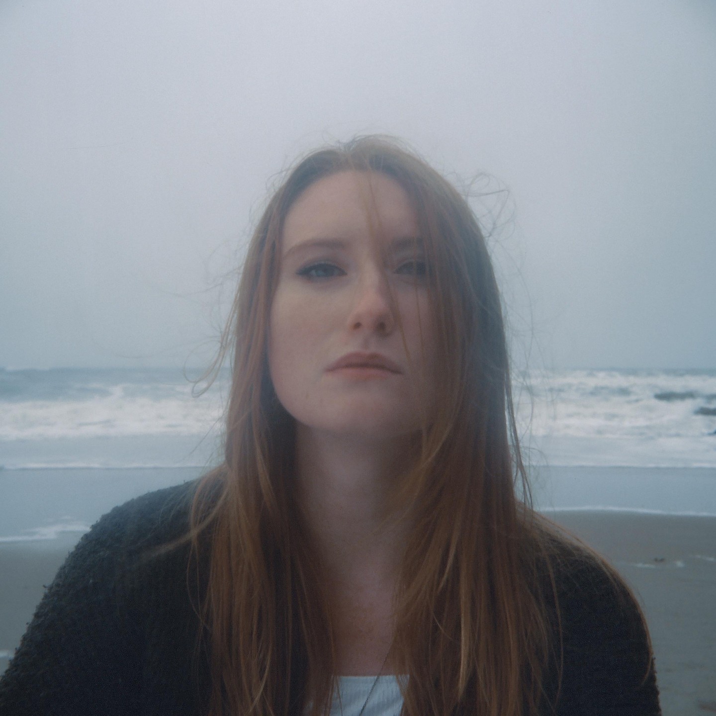 A woman with long hair stands on a beach with the ocean in the background.