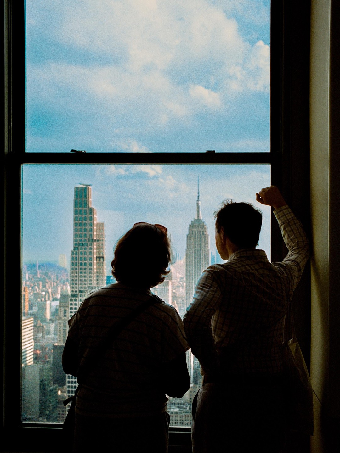 Two people stand silhouetted against a window, looking out at the New York City skyline with the Empire State Building visible.