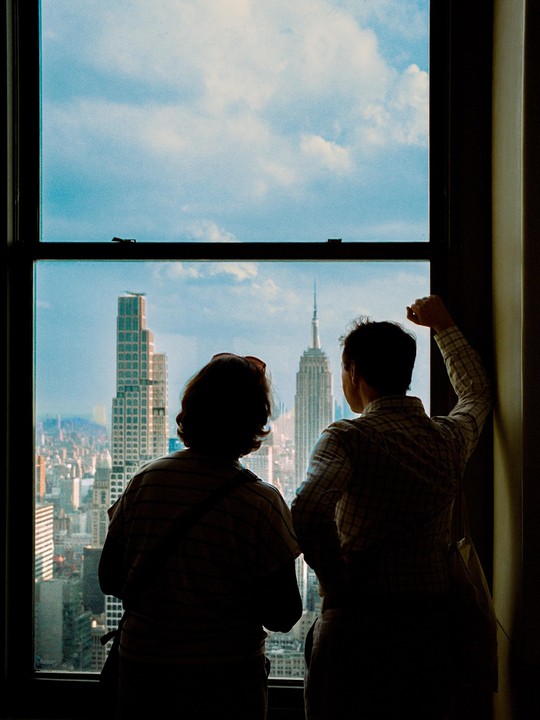Two people stand silhouetted against a window, looking out at the New York City skyline with the Empire State Building visible.