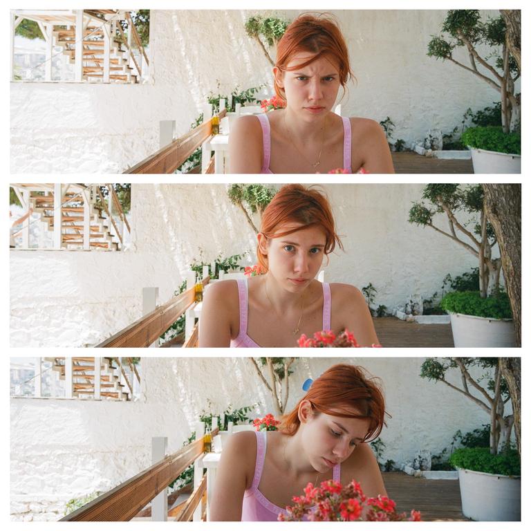 A woman with red hair sits at a table with red flowers in front of a white wall and a wooden railing.