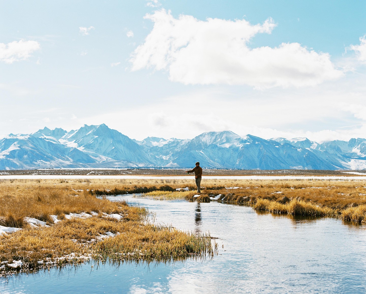 A man stands by a river with snow-capped mountains in the background.