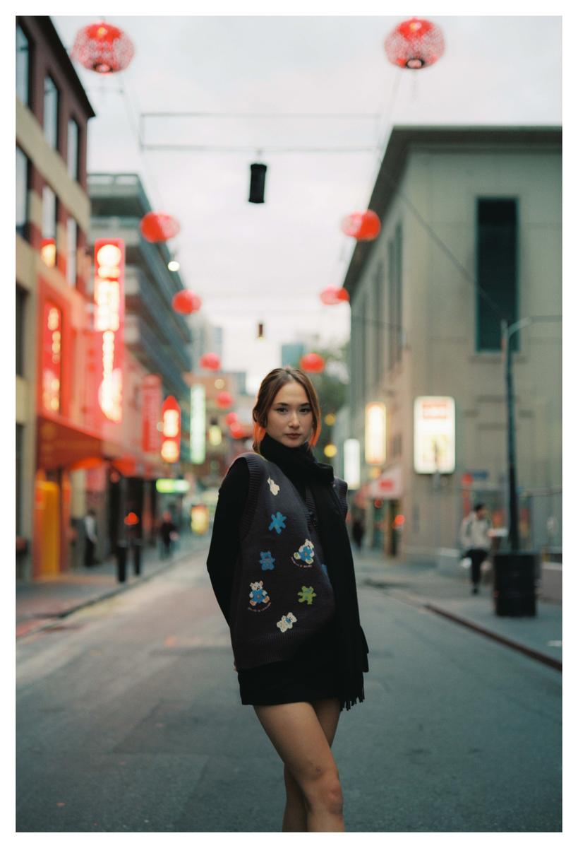 A woman stands in the middle of a street lined with buildings and red lanterns overhead.
