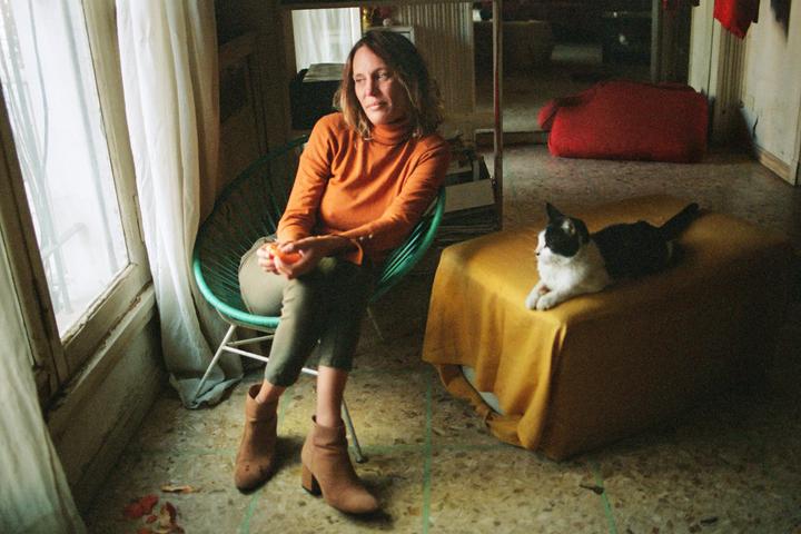 A woman sits in a chair next to a window while a cat lies on a yellow ottoman.
