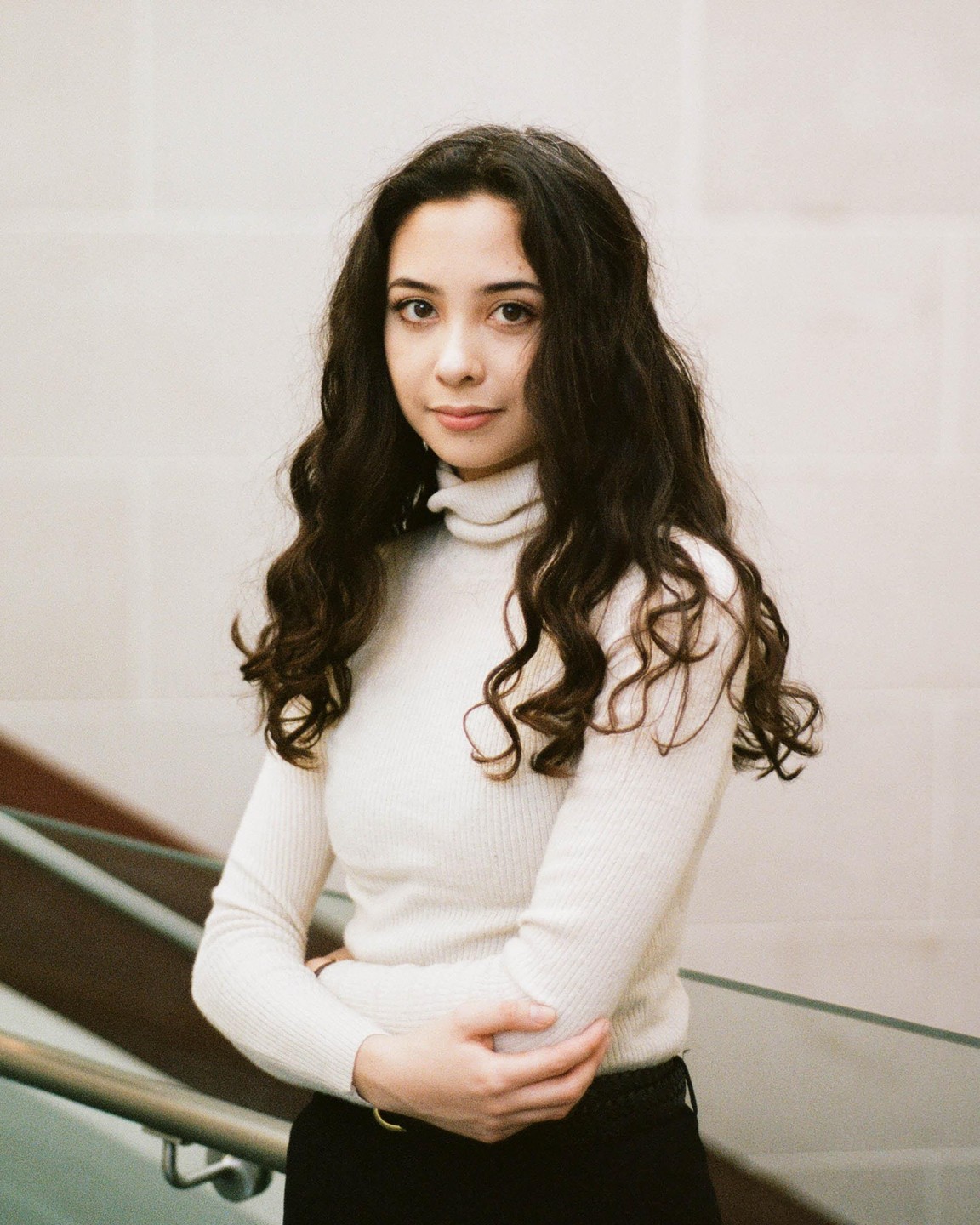 A woman with long, dark, curly hair wears a cream turtleneck and stands next to a railing.