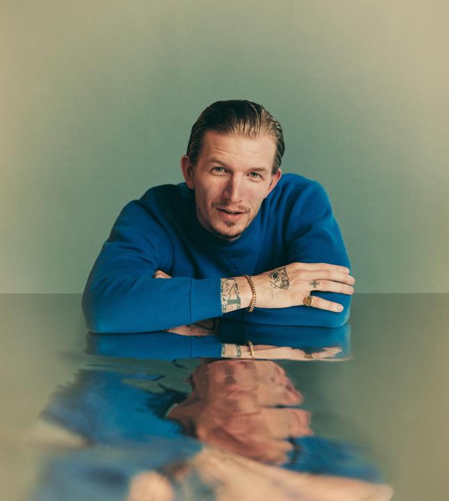 A man in a blue sweater sits at a table with his reflection visible in the surface.