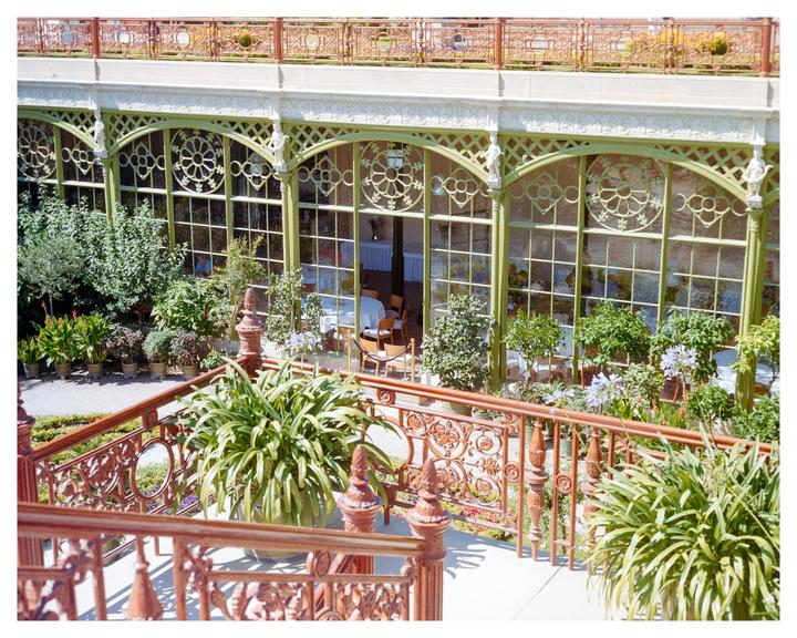 A grand conservatory with ornate green-framed windows and a balcony with a decorative railing is surrounded by lush potted plants.