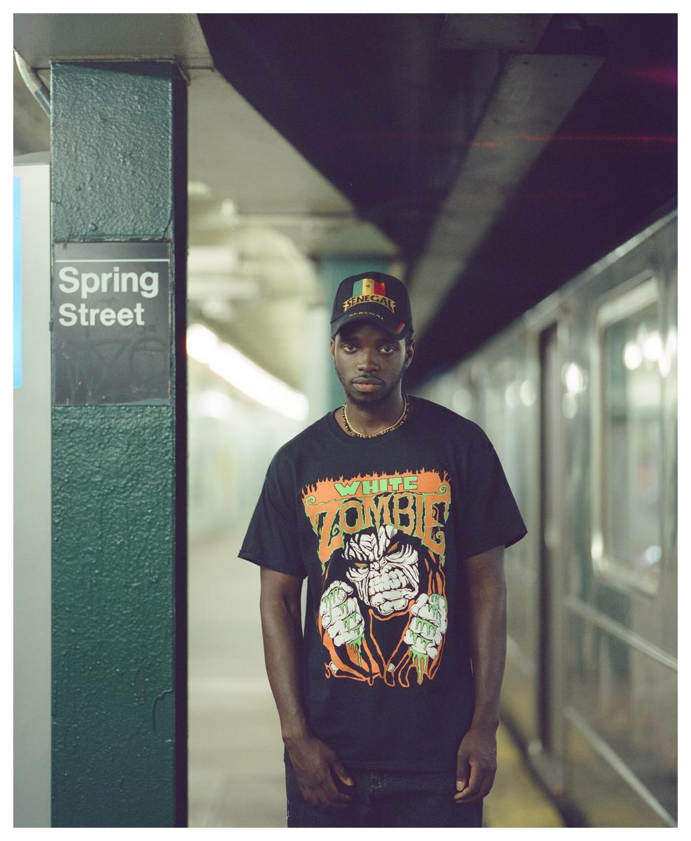 A man wearing a black t-shirt and a cap stands on a subway platform next to a sign that reads "Spring Street."