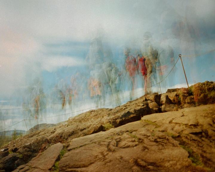 A rocky mountain slope is visible under a hazy sky with multiple exposures of people walking along a rope fence.
