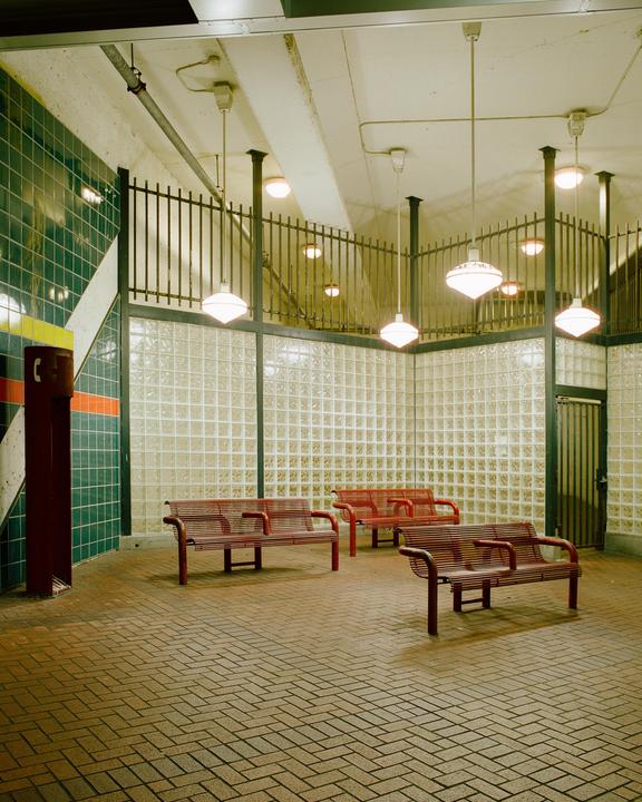 Three red benches sit on a brick floor in a waiting area with glass block walls and hanging lights.