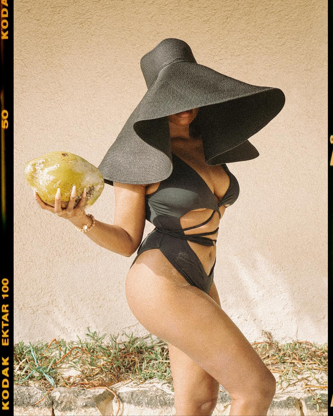 A woman in a black swimsuit and large hat holds a coconut.