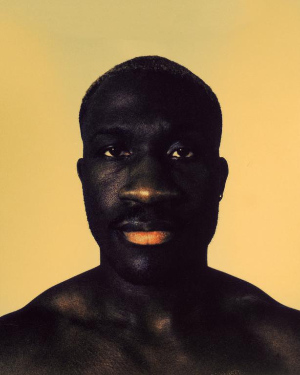 A man with dark skin and short hair looks directly at the viewer against a plain yellow background.
