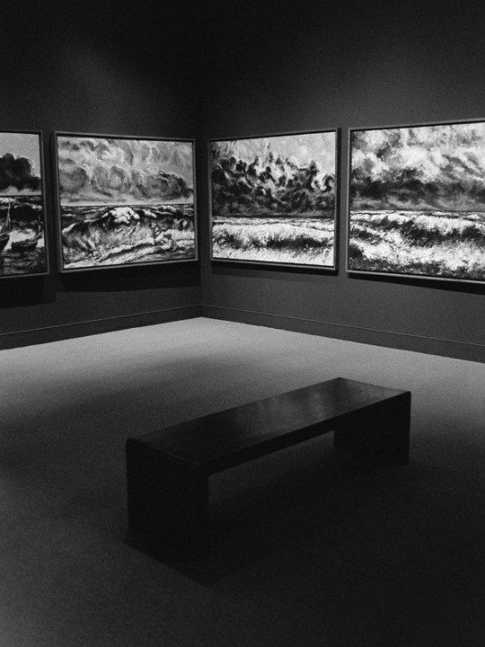 Four paintings of stormy seas hang on a dark wall above a bench in a gallery.