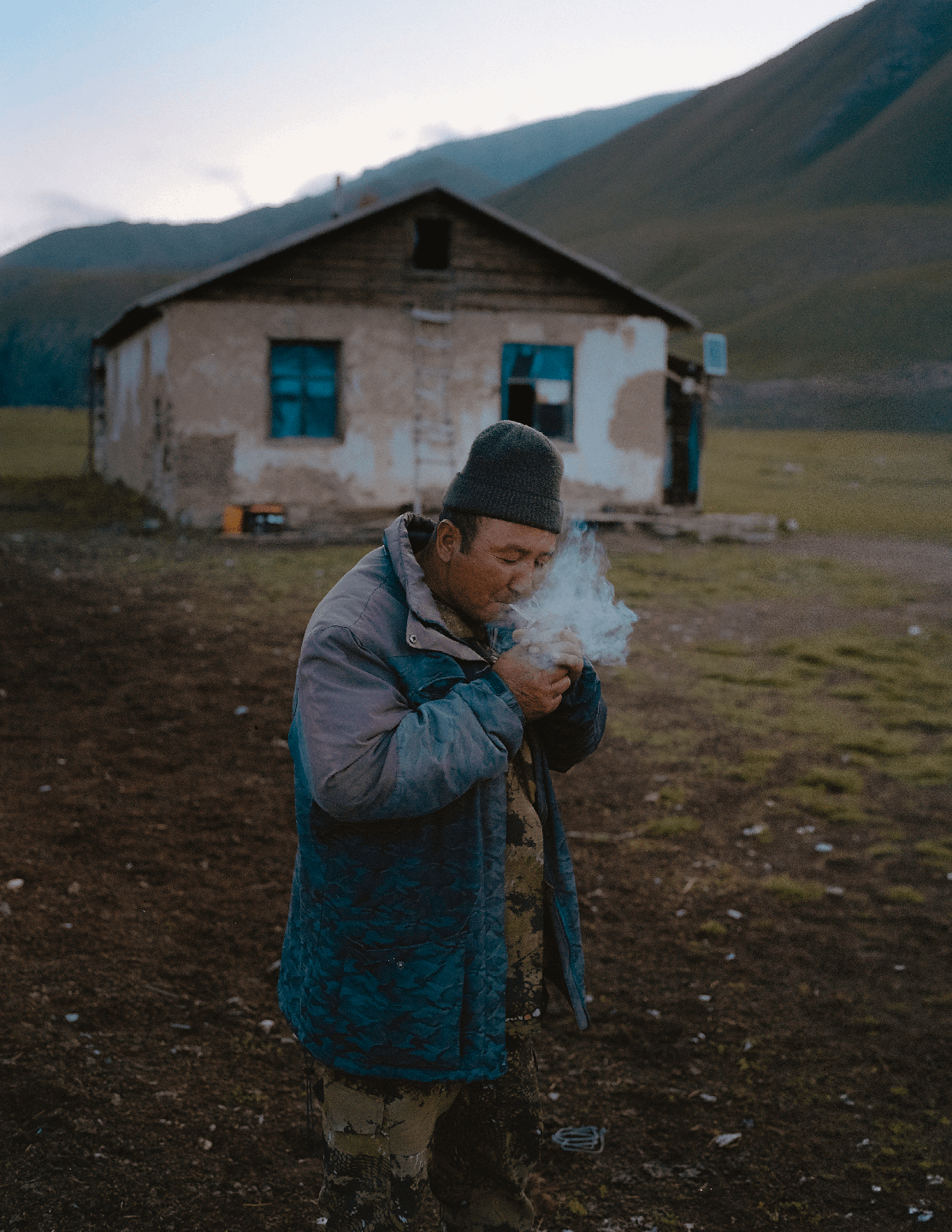 A man in a knit hat and jacket blows smoke from his mouth in front of a small house with mountains in the background.