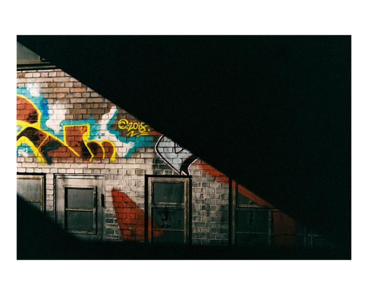 A brick wall with colorful graffiti and several windows is partially illuminated by a diagonal beam of light.