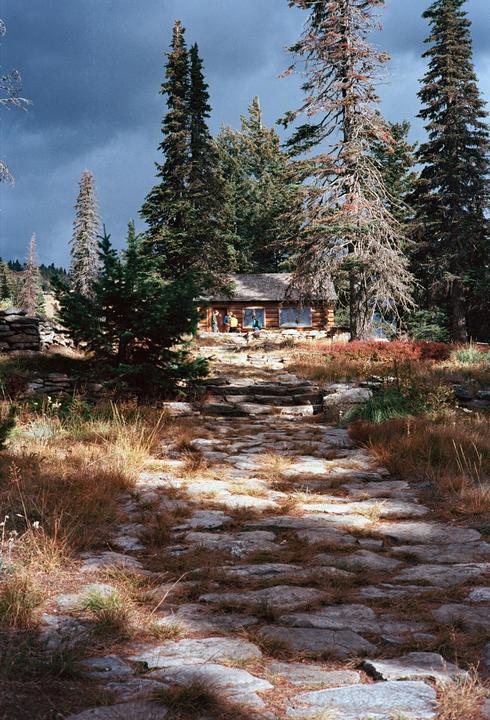 A stone path leads up to a wooden cabin surrounded by tall pine trees under a dramatic sky.