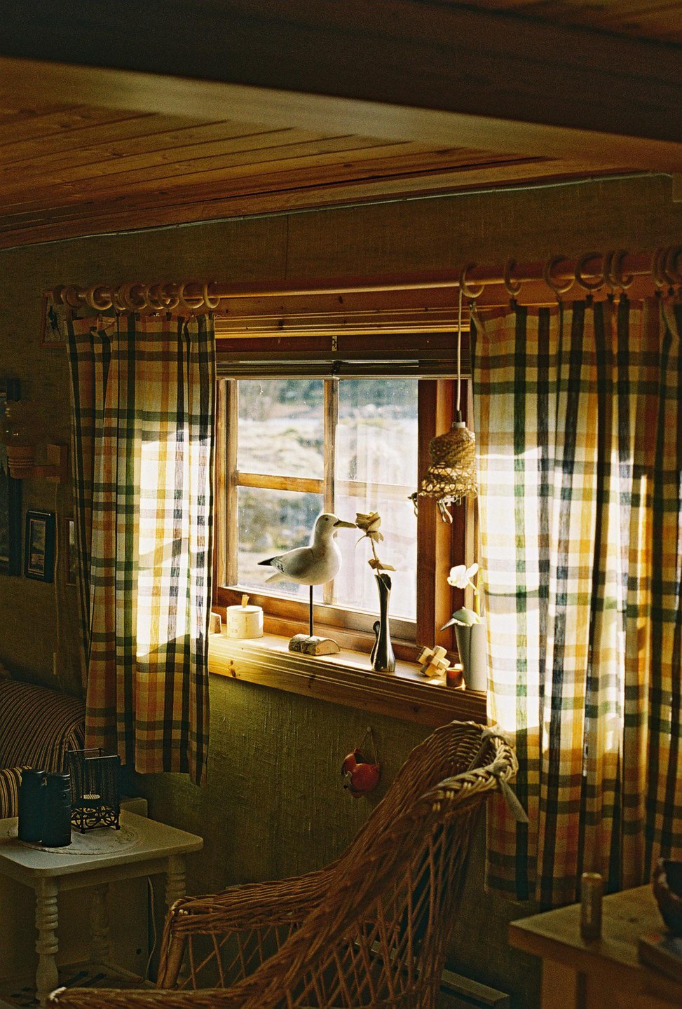 Sunlight streams through a window with plaid curtains, illuminating a wooden seagull figurine and other decorative items on the windowsill.