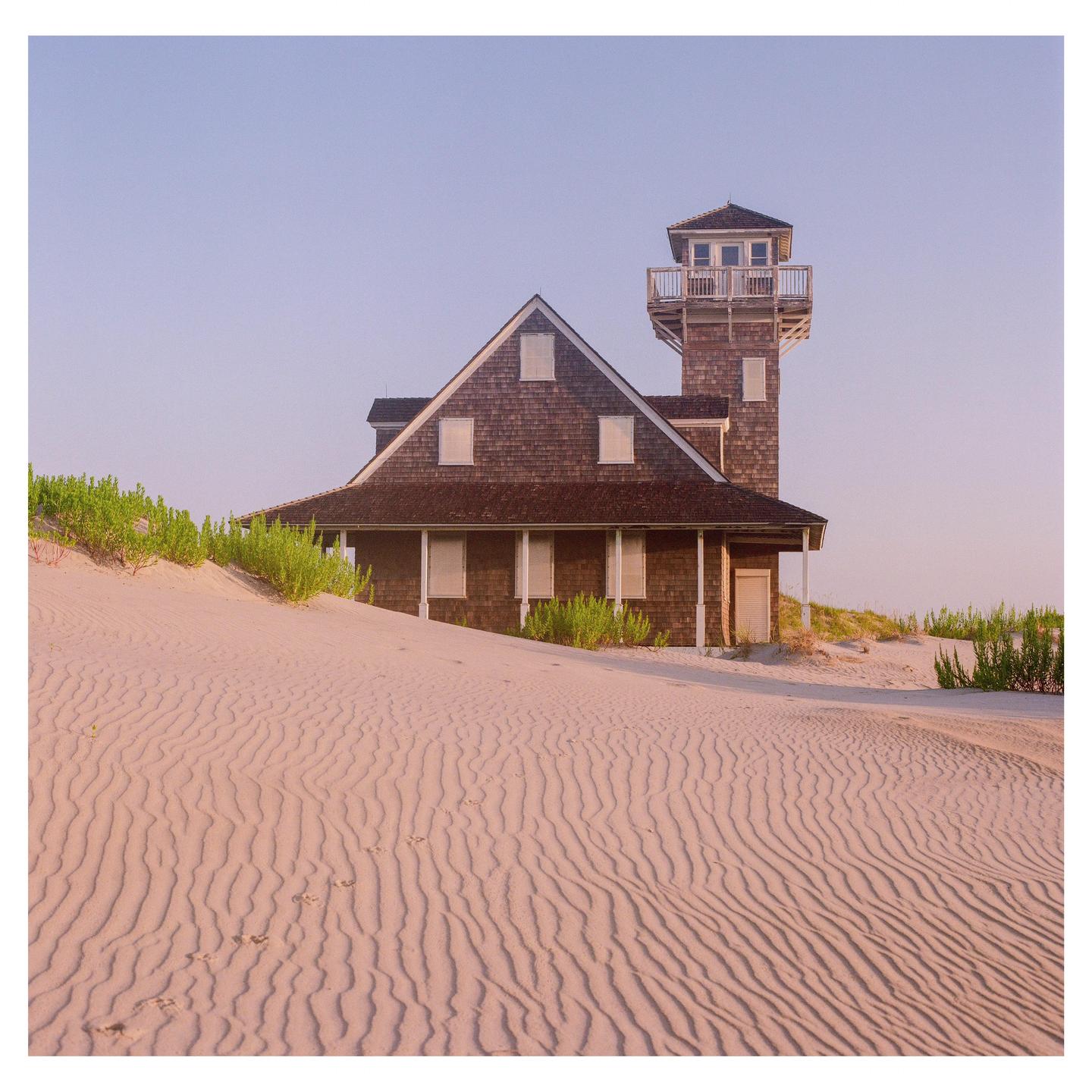 A wooden house with a tower stands on a sand dune with ripples in the foreground.