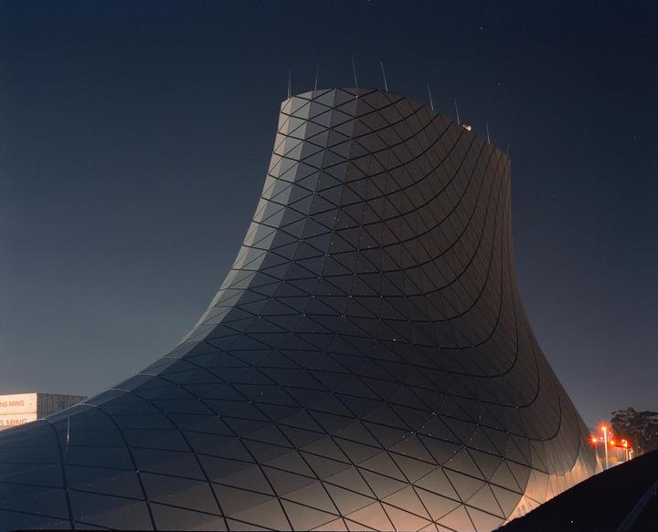 A modern building with a curved, gridded facade is illuminated at night under a dark sky.