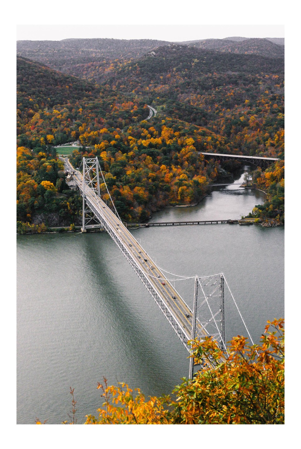 A suspension bridge with cars crosses a wide river, surrounded by hills covered in autumn foliage.