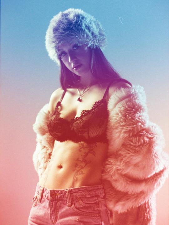 A woman wearing a fur hat, fur coat, and bra poses for a portrait.