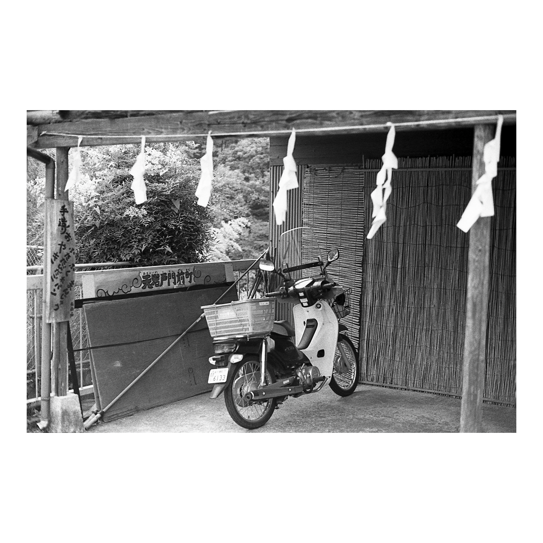 A scooter with a basket is parked under a wooden structure adorned with white paper strips, next to a bamboo screen.