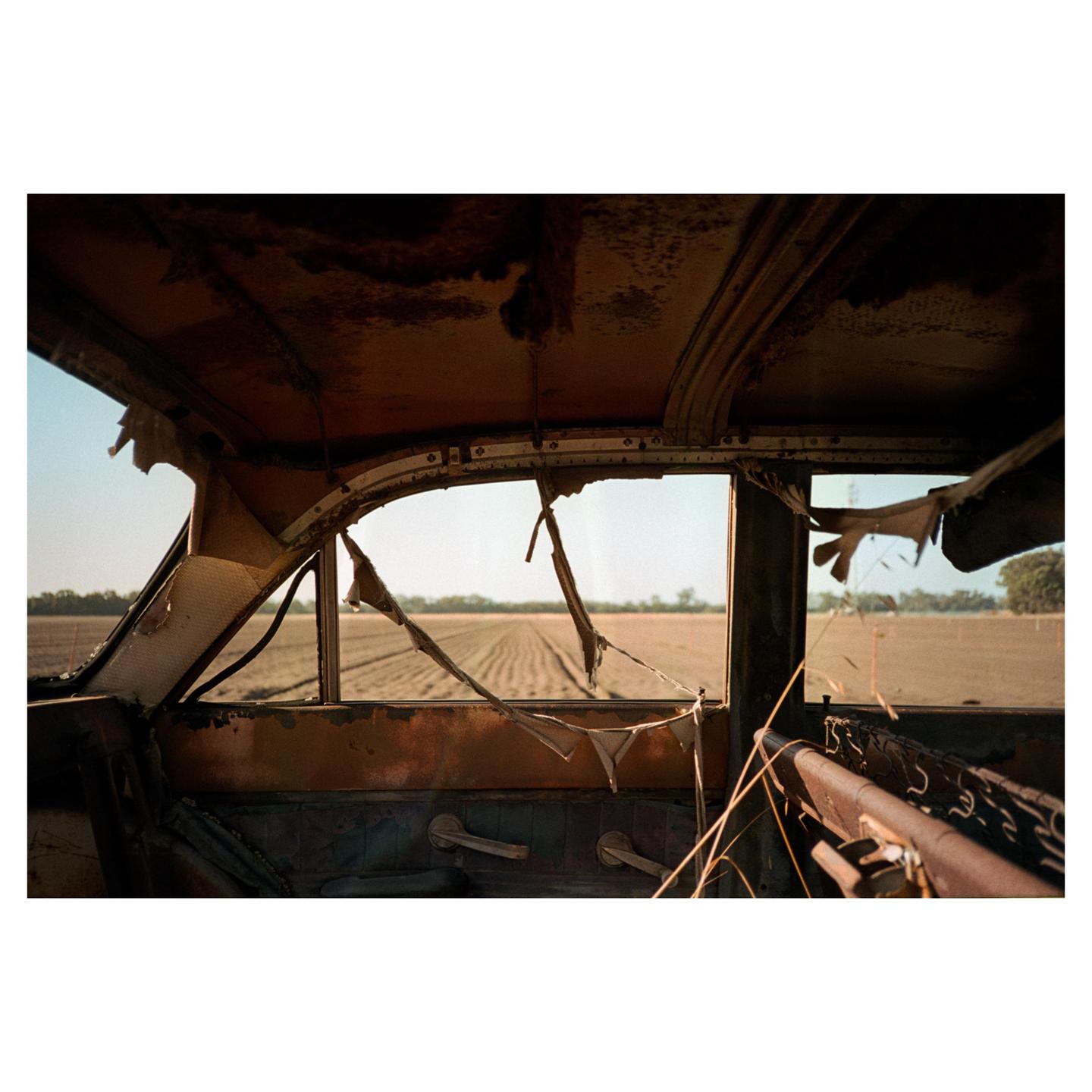 The interior of a rusted, broken-down vehicle looks out onto a field with rows of tilled earth.