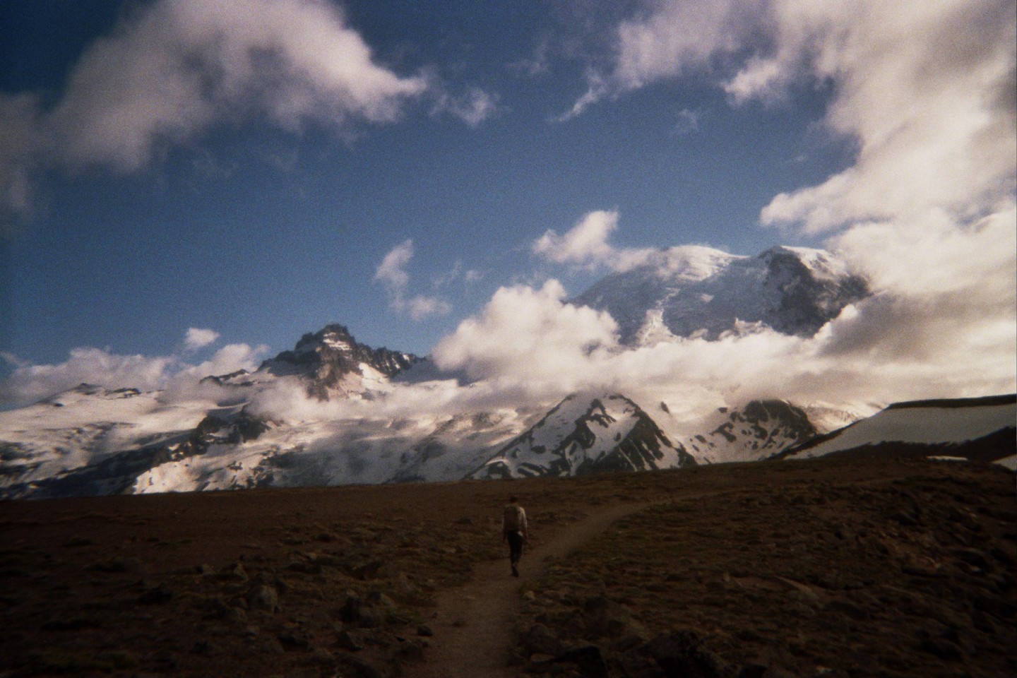 A person walks on a path towards snow-capped mountains partially obscured by clouds.