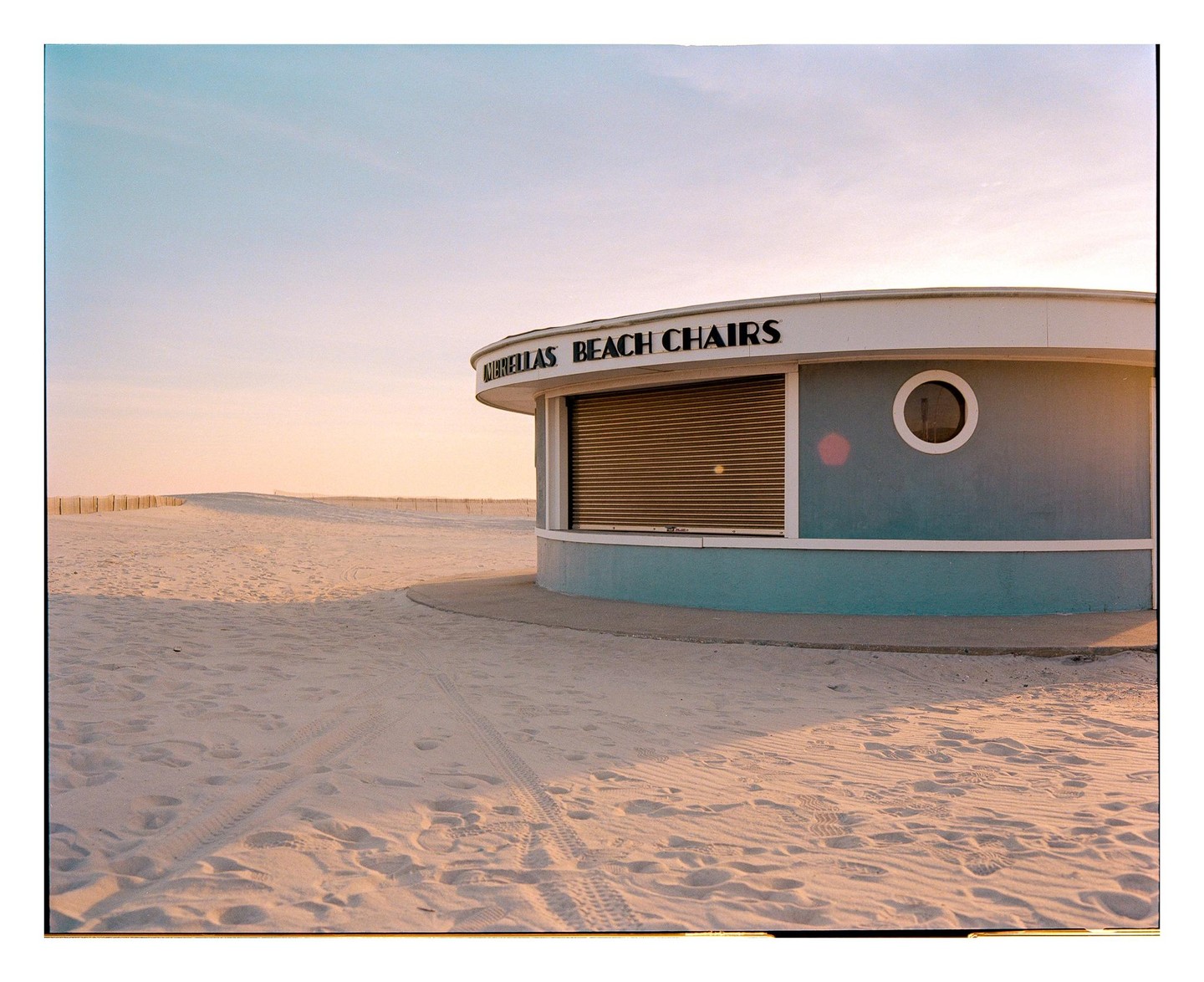 A closed beach chair rental stand with a circular window sits on a sandy beach under a clear sky.