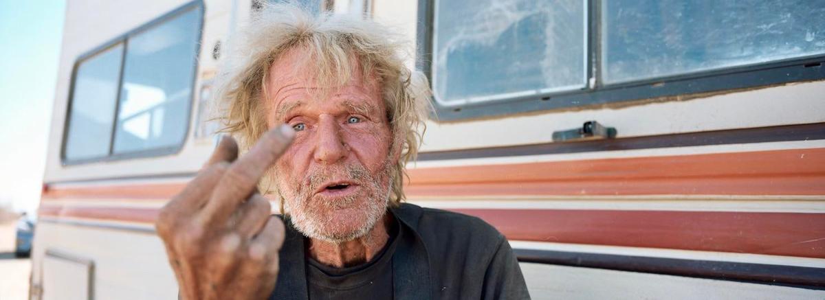 A man with messy hair and a beard gives the middle finger to the camera, with a recreational vehicle in the background.