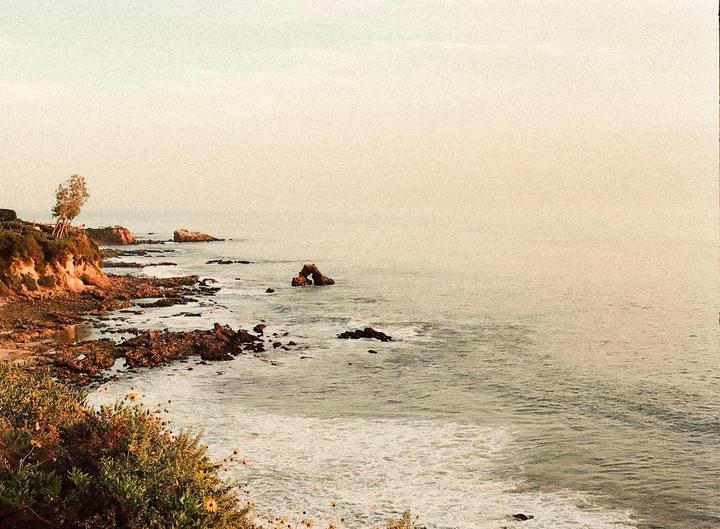 A hazy ocean view shows a rocky coastline with a natural arch rock formation in the water.