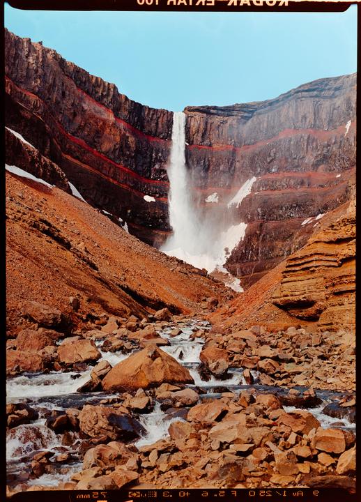 A powerful waterfall cascades down a red and brown rocky cliff into a riverbed filled with rocks and flowing water.