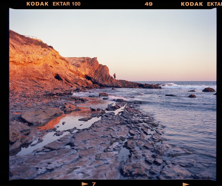 A man stands on a rocky outcrop at the edge of the ocean, with cliffs illuminated by the setting sun.