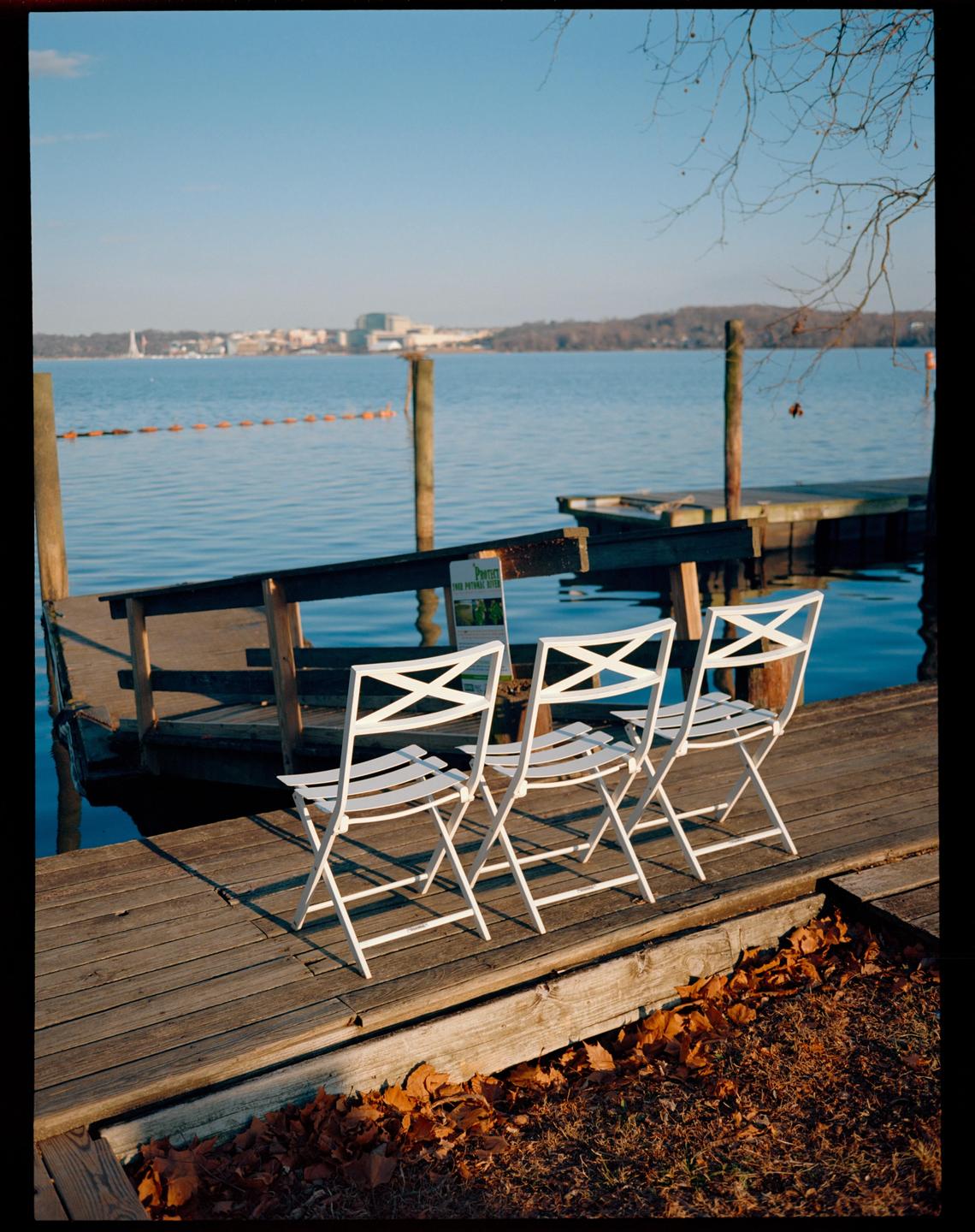 Three white folding chairs sit on a wooden dock next to a body of water with a distant shoreline.