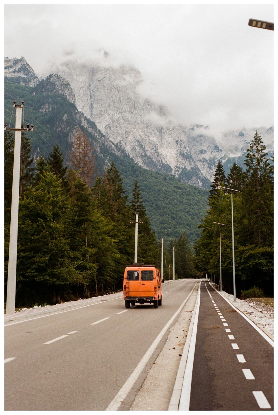An orange van drives on a road through a forest with a large, cloudy mountain in the background.