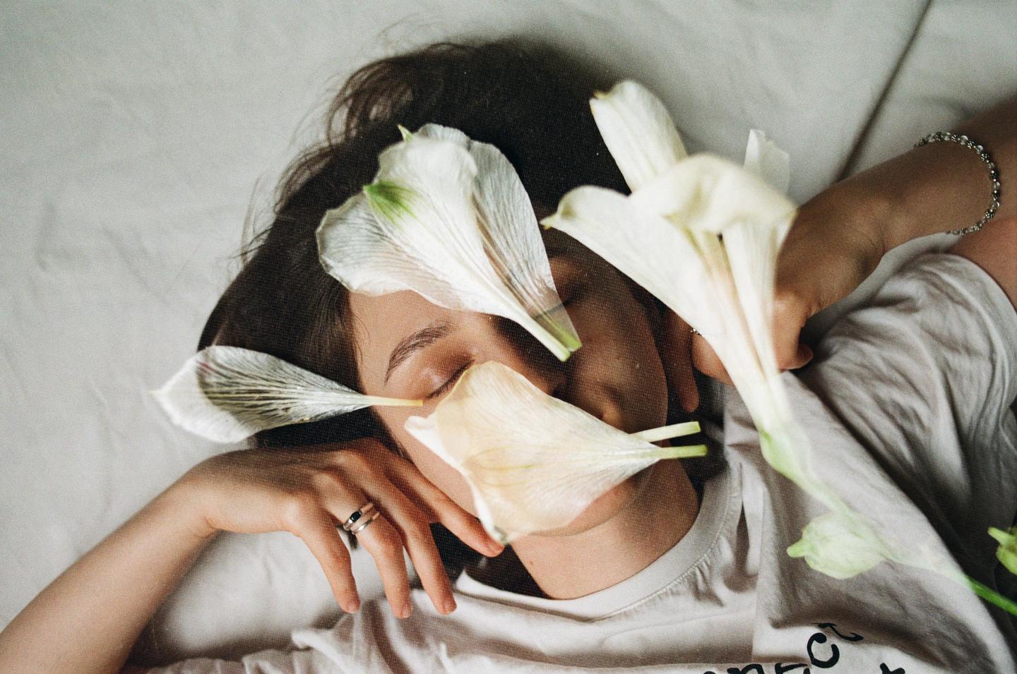 A woman lies with her eyes closed, covered in white flower petals.