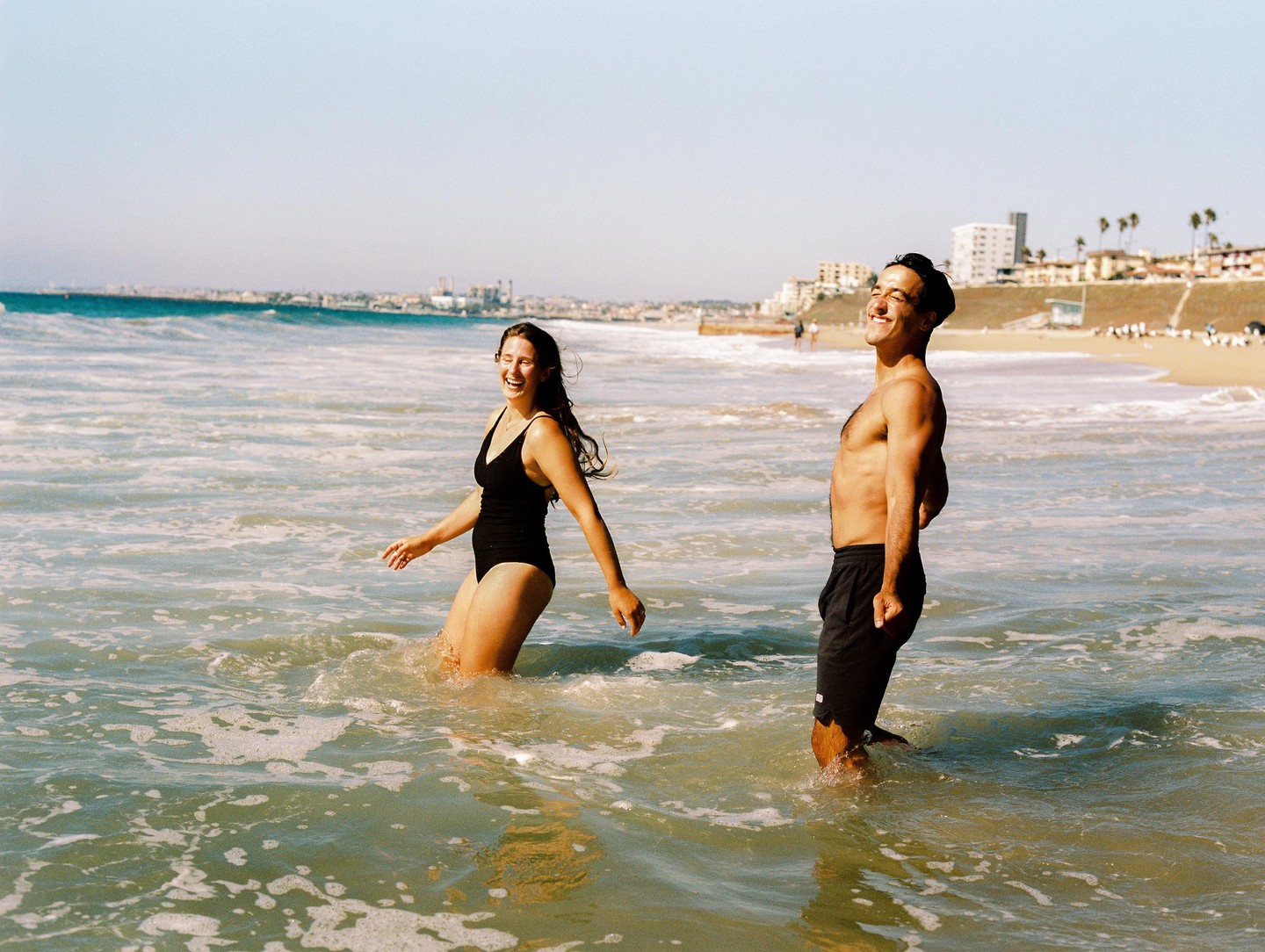 A couple stands in the ocean with a beach and buildings in the background.