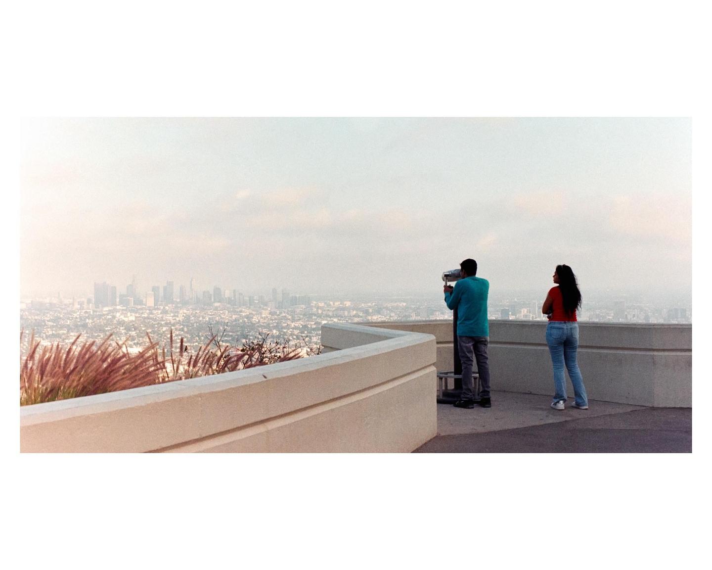 A man looks through a coin-operated telescope while a woman stands beside him, overlooking a hazy city skyline.