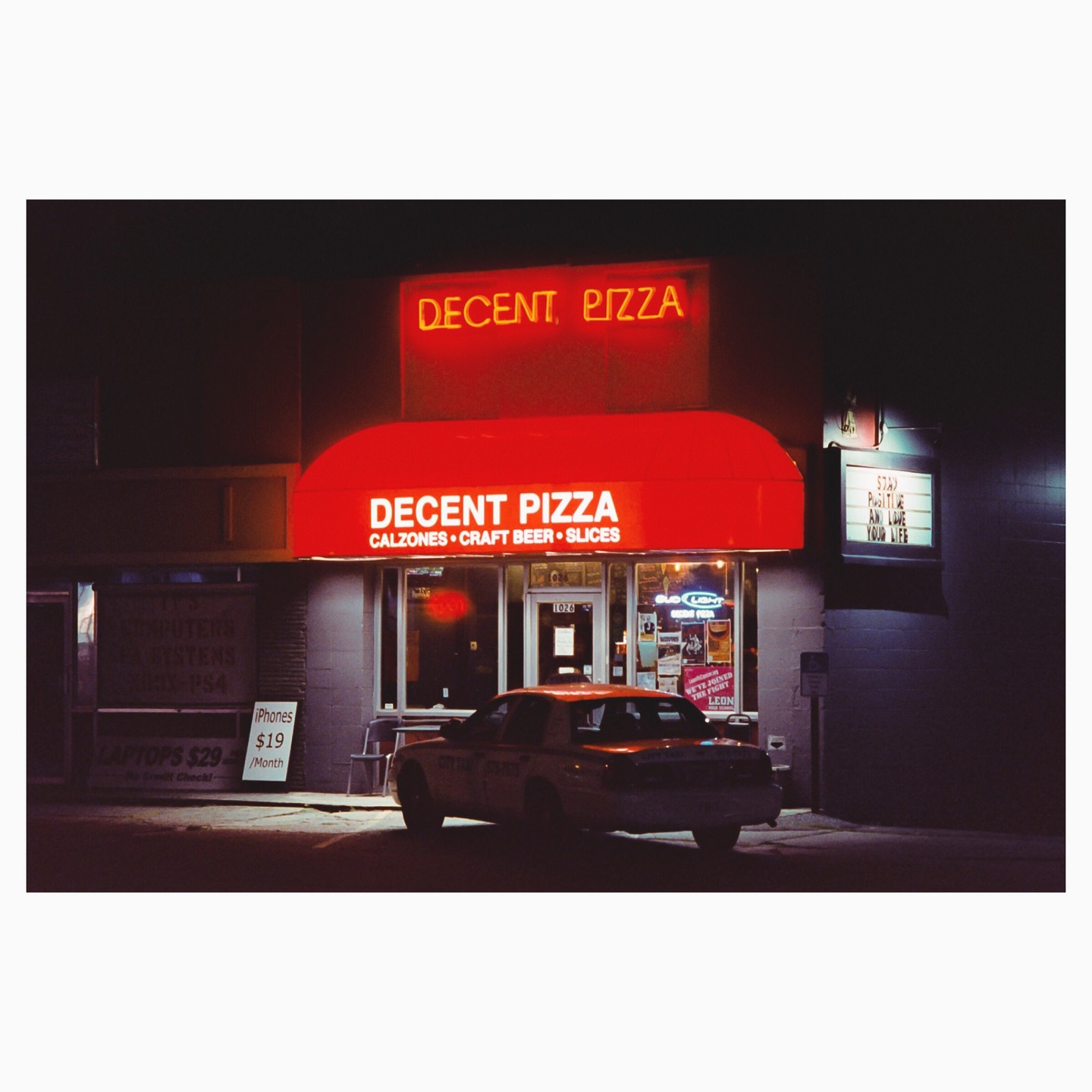 A car is parked in front of a pizza shop with a red awning and neon signs at night.