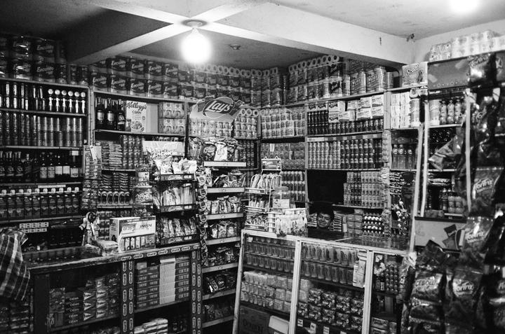 A man smiles from behind a counter in a small store filled with shelves of products.