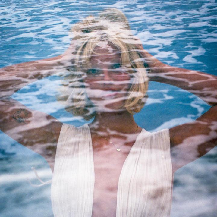 A woman with blonde hair and a white dress is visible through a double exposure with blue water.