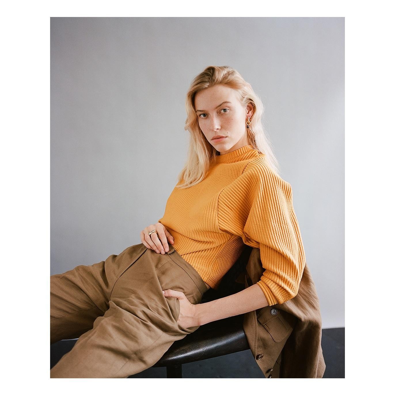 A woman with long blonde hair and an orange top sits on a chair with her hand in her pocket.