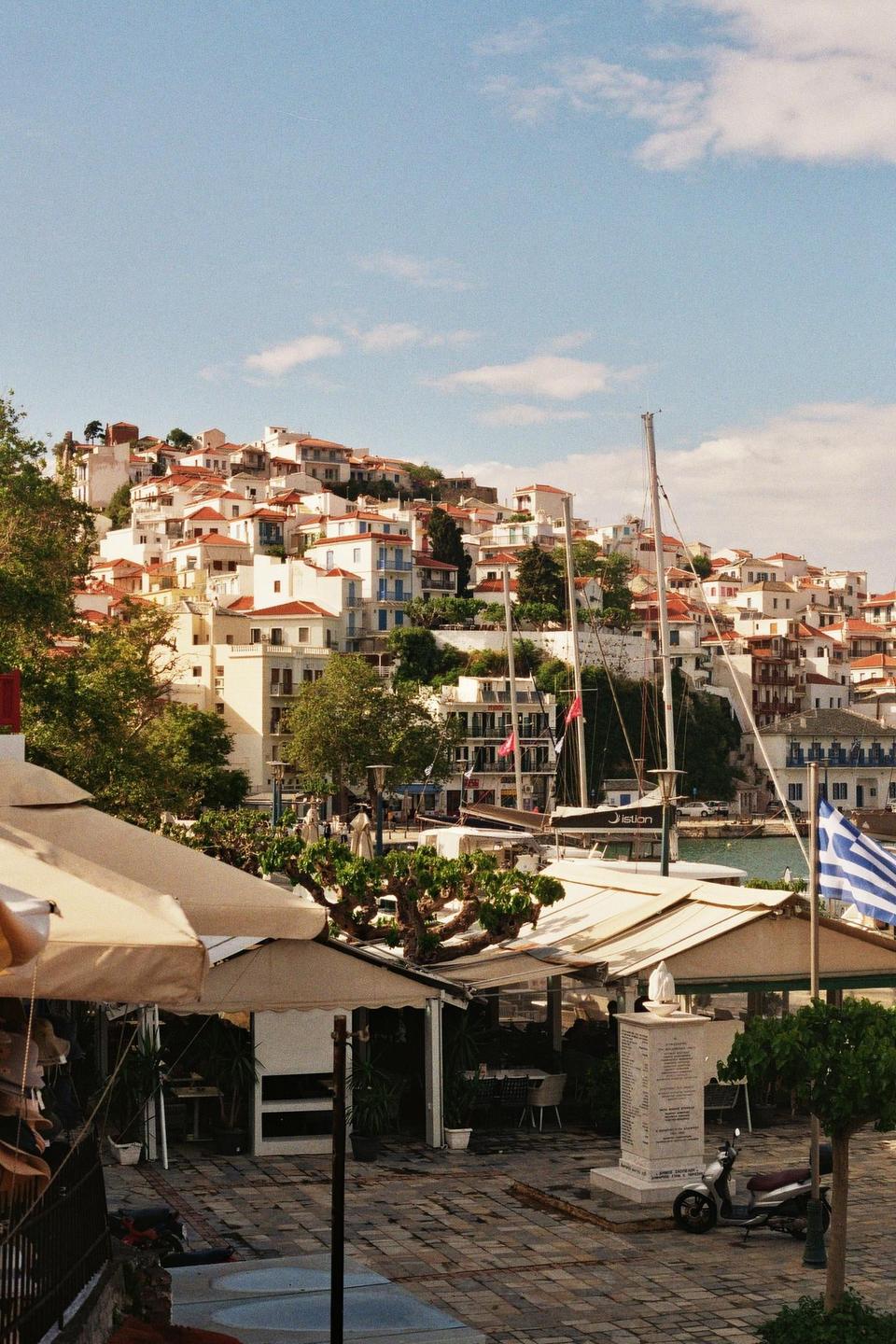 A hillside town with white buildings and red roofs overlooks a harbor with boats and a Greek flag.