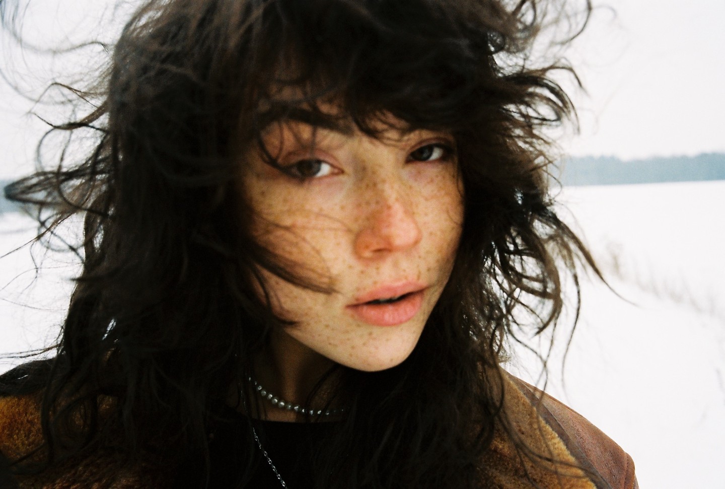 A woman with freckles and dark, windswept hair looks directly at the viewer in a snowy landscape.